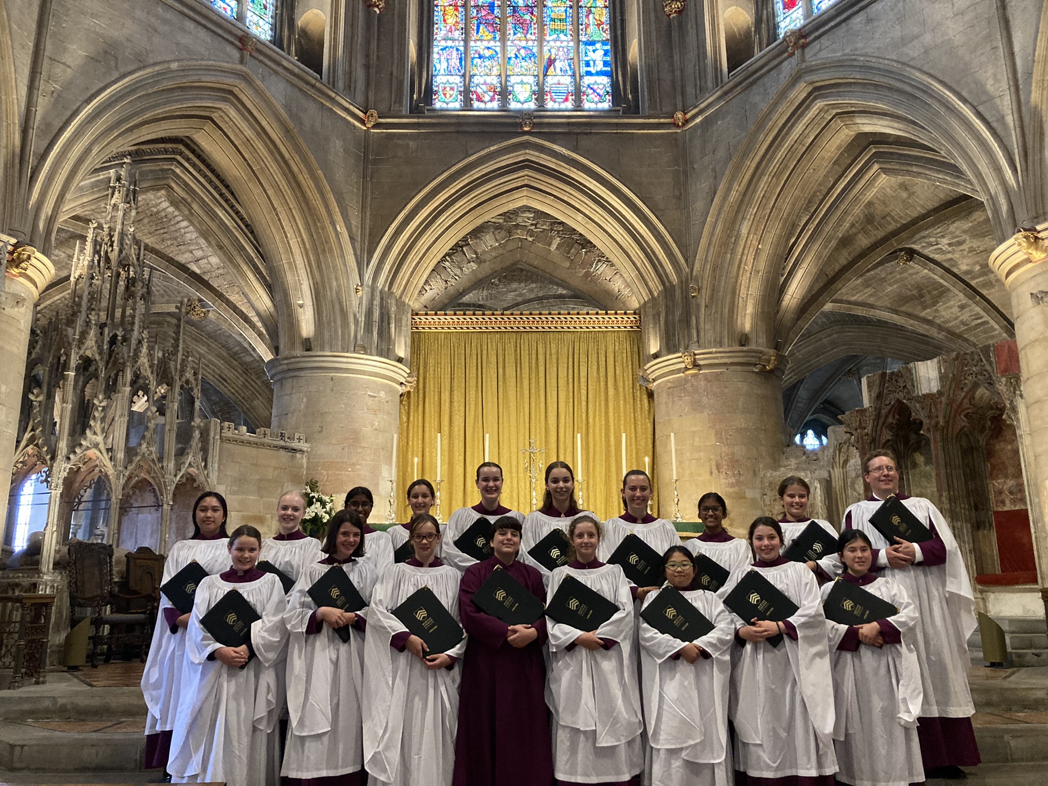 A group of Choristers in burgundy cassocks and white surplices stand in Tewkesbury Abbey holding their black and gold music folders