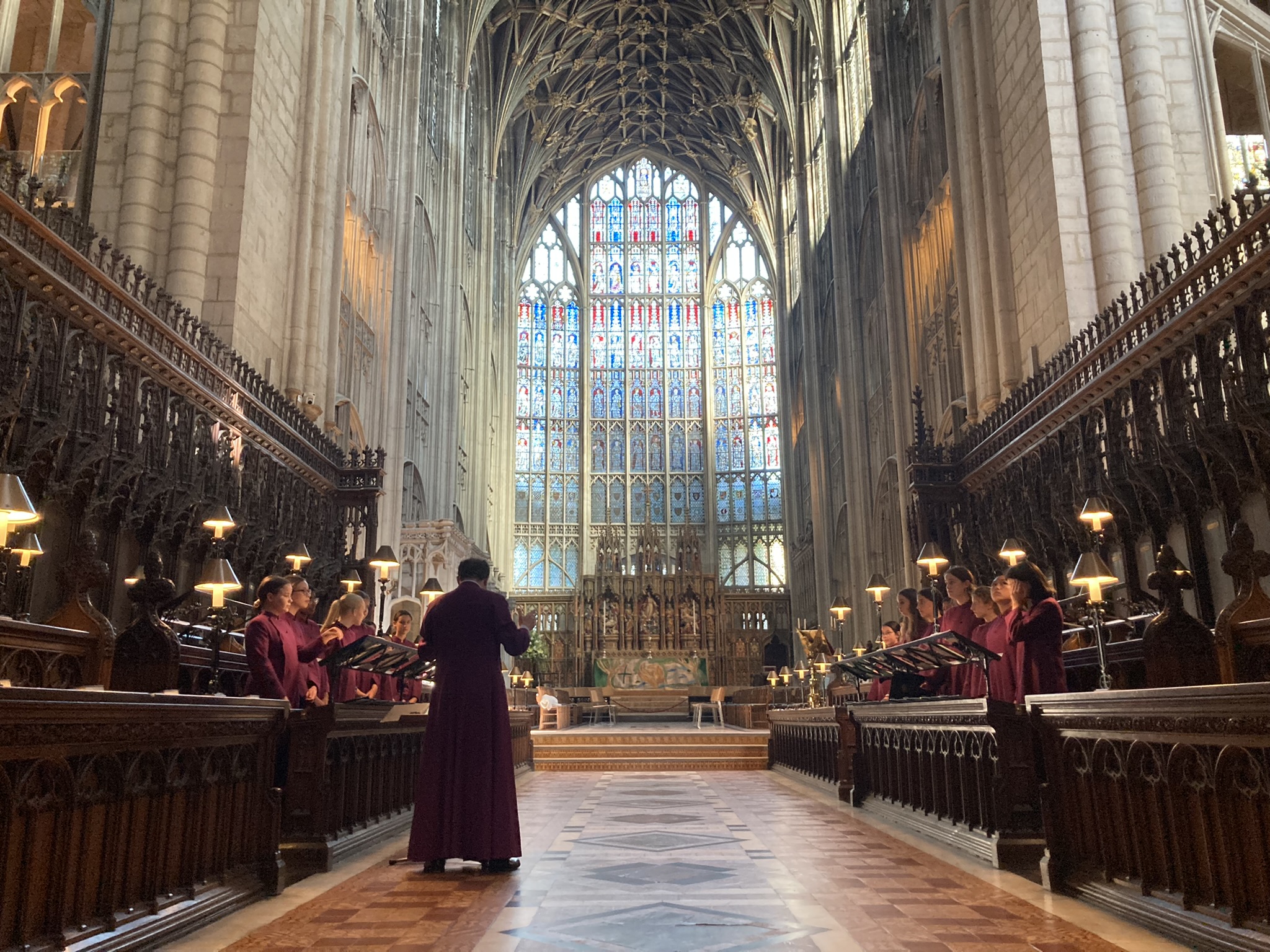 The choristers sing in their burgundy cassocks in the stalls in front of the large blue and red glass window of of Gloucester Cathedral