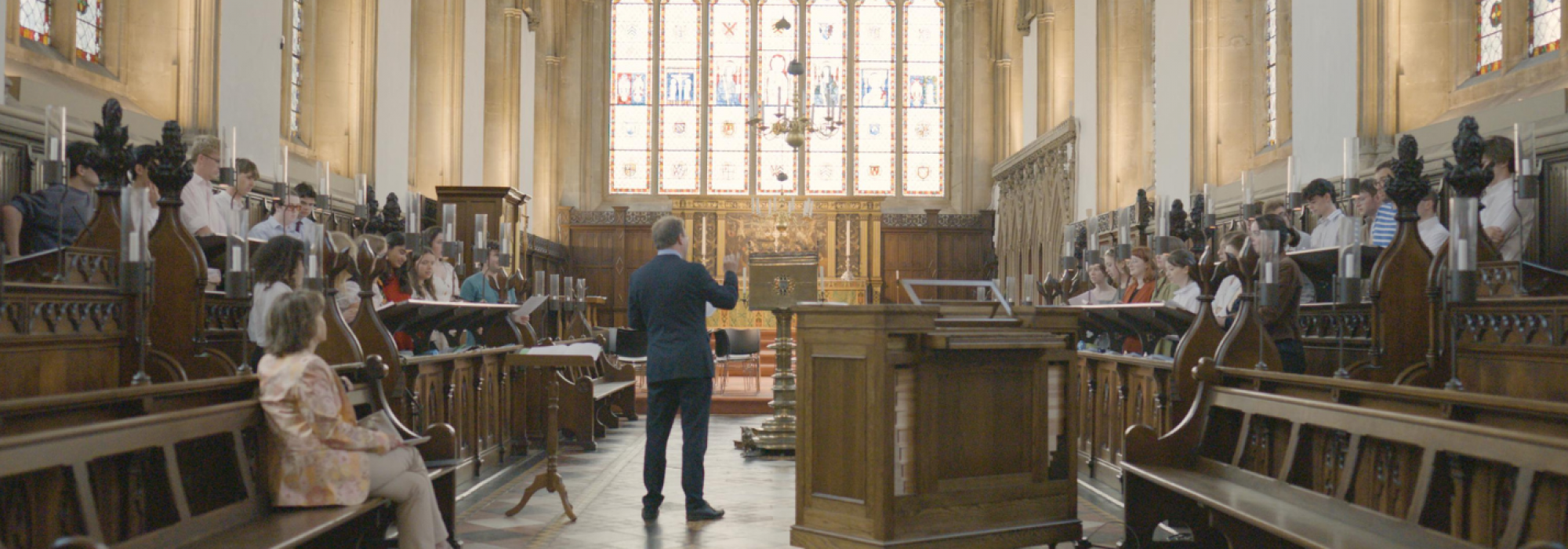 A wide span of the Chapel facing the stained glass at the East end with the choir in stalls rehearsing, the Director of Music conducting in a blue suit, and composer Roxanna Panufnik observing