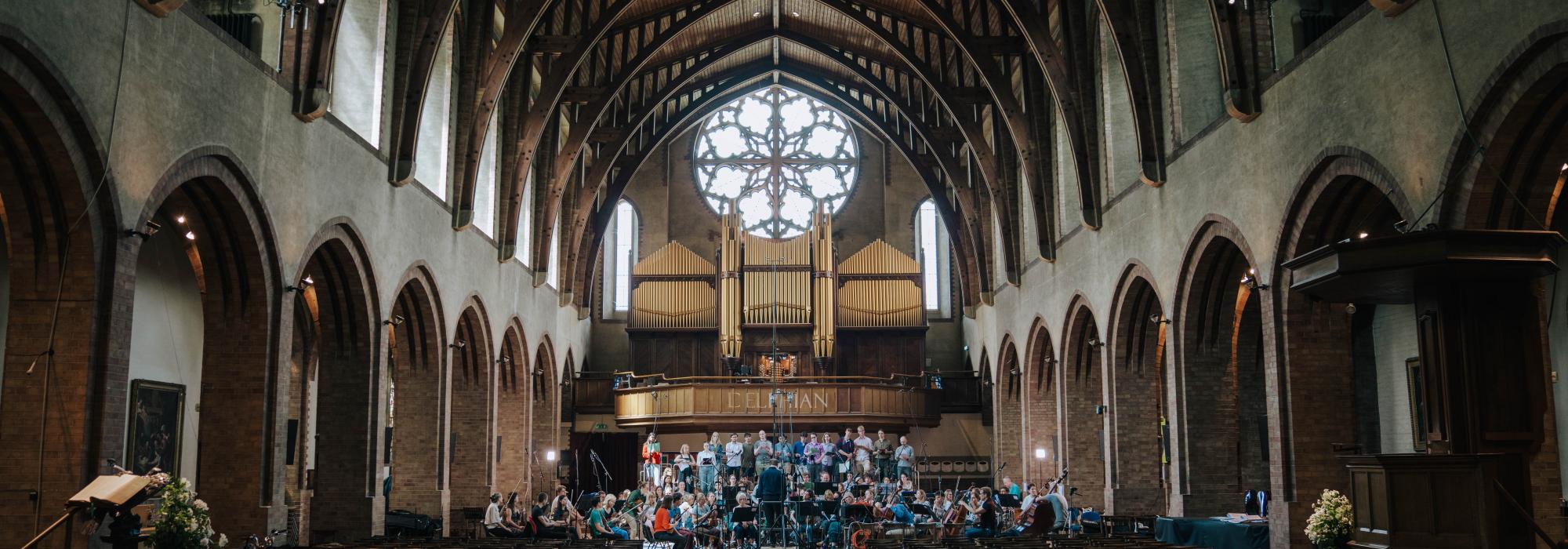 The choir and orchestra in dimly lit chapel surrounded by microphones