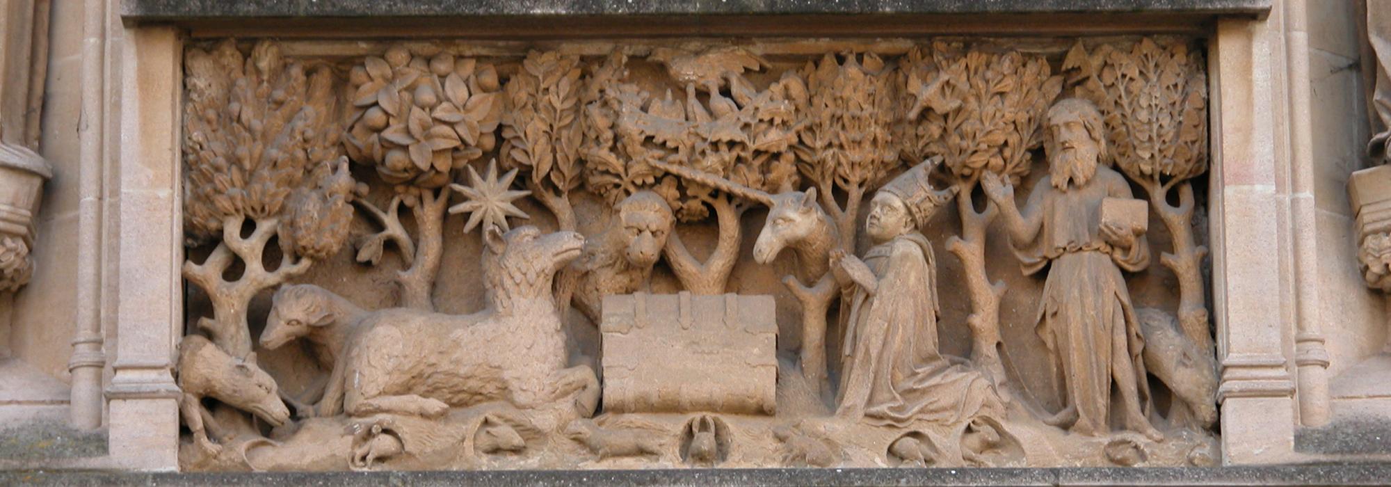 Central stone panel above the entrance to the College; it represents a book with the Agnus Dei on one side and St John the Baptist and the founder on the other side set in a landscape background with various animals. Central stone panel above the entrance to the College; it represents a book with the Agnus Dei on one side and St John the Baptist and the founder on the other side set in a landscape background with various animals.