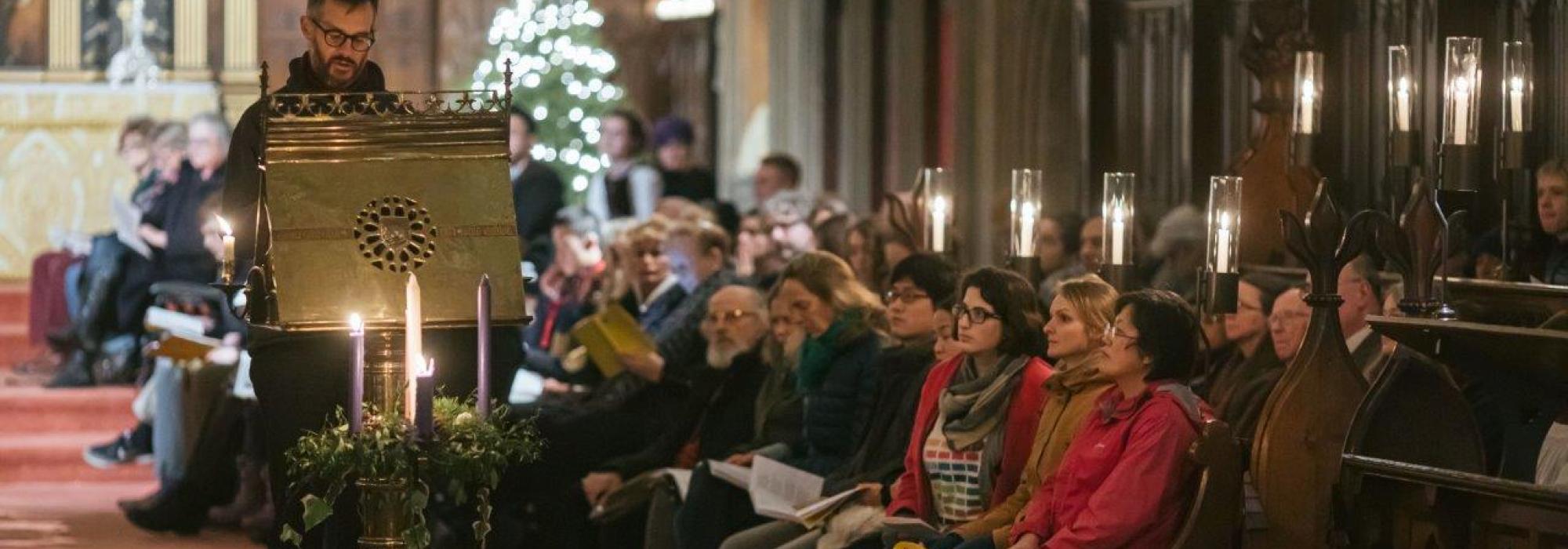 Christmas Carol Service in Chapel lit by candles with Christmas trees in the background. A man reads a lesson from the lectern. 