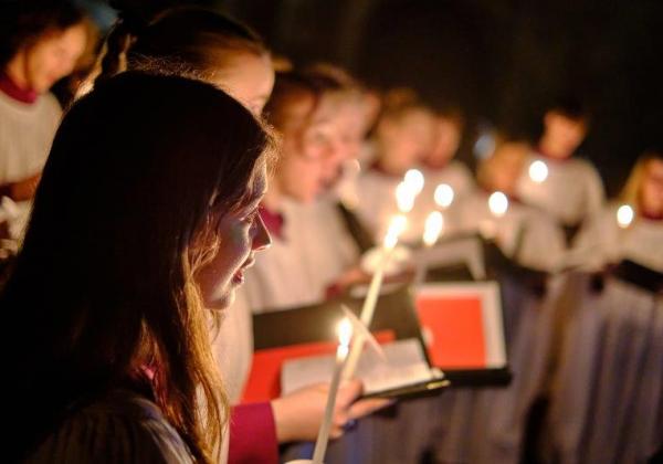 Choir members in soft focus with red choir books hold candles in the dimly lit antechapel
