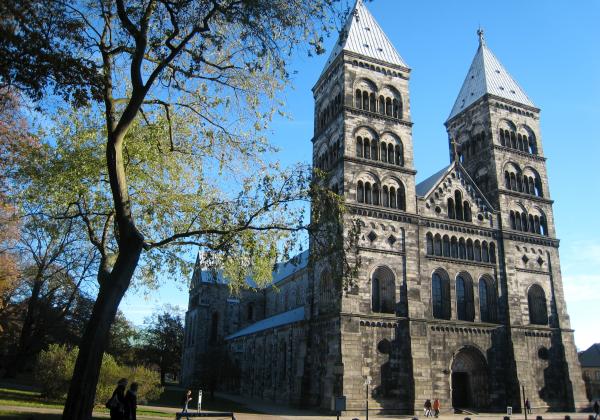 Two grey stone towers of Lund Cathedral with a tree in the foreground and a blue sky