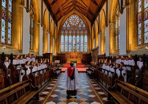 The choir stand in the stalls, conducted by Ben Nicholas in robes, in front of the Merton Chapel East windows