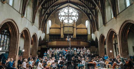 Choir and full orchestra surrounded by microphones and lit by a large church window