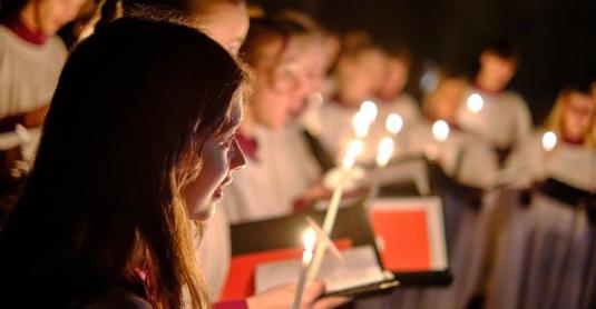 Choir members in soft focus with red choir books hold candles in the dimly lit antechapel