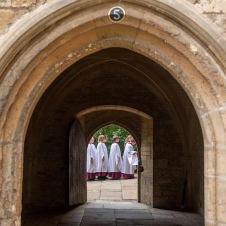 A line of girl choristers in white surplices are viewed through a stone archway 
