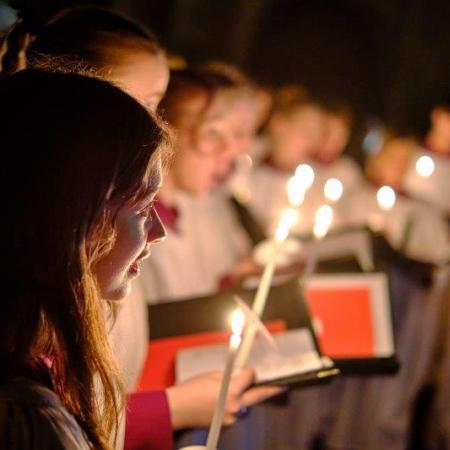 Choir members in soft focus with red choir books hold candles in the dimly lit antechapel